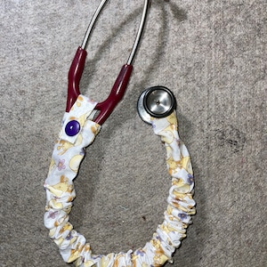 May include: A stethoscope with a white fabric cover featuring a pattern of yellow bunnies and purple flowers. The stethoscope has a burgundy coloured tubing and a silver metal head.