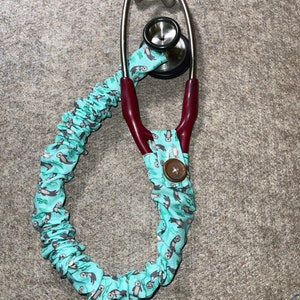 Peut inclure: Un stéthoscope avec un revêtement en tissu bleu sarcelle orné d'un motif de loutres de mer. Le stéthoscope a un tube de couleur bordeaux et un embout thoracique argenté.