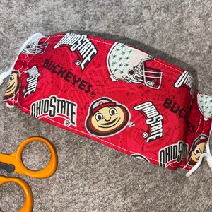 May include: Red fabric face mask with a white drawstring. The mask has a repeating pattern of the Ohio State logo, the word "Buckeyes", and a cartoon mascot.
