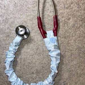 Peut inclure: Un stéthoscope avec un revêtement en tissu à motif bleu clair. Le stéthoscope a un cadre en métal argenté et un tube en caoutchouc rouge.