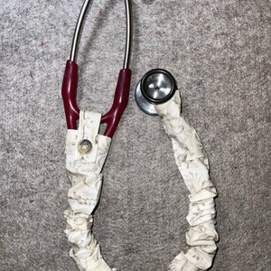 Peut inclure: Un stéthoscope avec un revêtement en tissu floral blanc. Le stéthoscope a un tube rouge et argenté et un pavillon argenté.