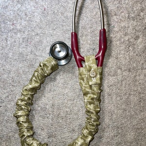 Peut inclure: Un stéthoscope rouge avec un revêtement en tissu vert à motif de libellule. Le stéthoscope a une seule tête et un tube double.