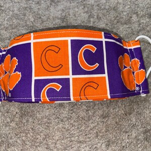 May include: Purple and orange face mask with a checkered pattern featuring the letter "C" and paw prints.