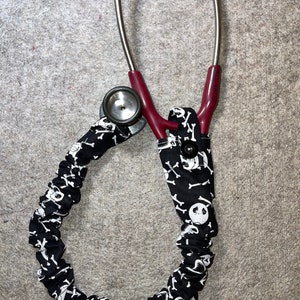 Peut inclure: Un stéthoscope rouge avec un revêtement en tissu noir et blanc à motif de crânes et d'os croisés. Le stéthoscope est enroulé autour du revêtement en tissu.
