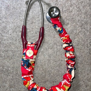 May include: A stethoscope with a red and white Wonder Woman fabric cover. The cover features a repeating pattern of the superheroine, stars, and logos. The stethoscope has a button closure and a silver chest piece.