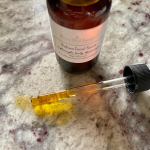 May include: A glass bottle with a dropper filled with a light amber liquid. The bottle has a white label with the text "Bloom Wild Beauty Radiant Facial Serum Lightweight Daily Moisturizer Made in Victoria, BC bloomwildbeauty.com".