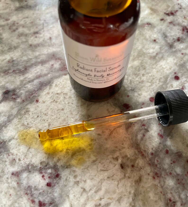 May include: A glass bottle with a dropper filled with a light amber liquid. The bottle has a white label with the text "Bloom Wild Beauty Radiant Facial Serum Lightweight Daily Moisturizer Made in Victoria, BC bloomwildbeauty.com".