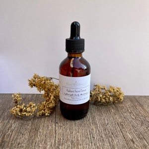 May include: A brown glass bottle with a dropper top, labeled "Radiant Facial Serum Lightweight Daily Moisturizer" by Bloom Wild Botanicals. The bottle is sitting on a wooden surface with dried flowers.
