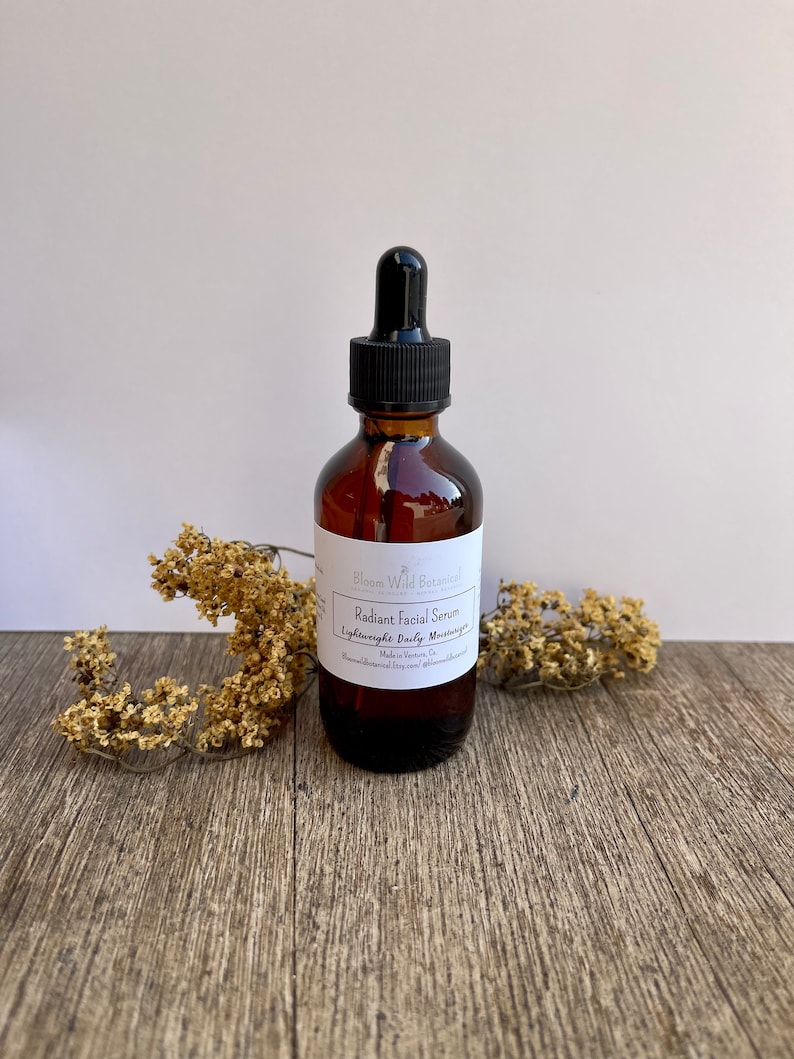 May include: A brown glass bottle with a dropper top, labeled "Radiant Facial Serum Lightweight Daily Moisturizer" by Bloom Wild Botanicals. The bottle is sitting on a wooden surface with dried flowers.