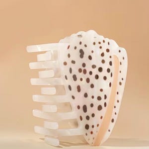 Puede incluir: Una pinza para el pelo grande, blanca y con manchas marrones, con forma de concha. La pinza tiene una serie de dientes rectangulares blancos en un lado. El diseño de la concha presenta manchas marrones y una franja de color melocotón claro.