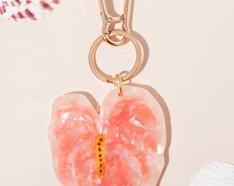 May include: A keychain featuring a heart-shaped, translucent resin charm with a marbled peach and white pattern. The charm includes a central yellow spadix detail with small red dots. It is attached to a gold-toned metal lobster clasp and ring.