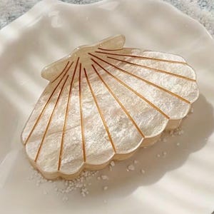 Puede incluir: Una pinza para el pelo con forma de concha nacarada con detalles dorados. La pinza descansa sobre un plato blanco con borde festoneado. El diseño de la pinza presenta líneas radiales, imitando la estructura natural de una concha.