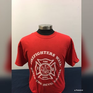 May include: Red t-shirt with a white graphic of a firefighter's helmet and crossed axes with the text "Firefighters Walk Where The Devil Dances" and "Fire Dept."