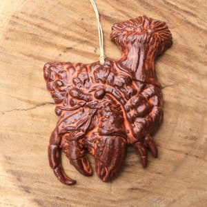 May include: A brown ceramic ornament shaped like a crab with a textured surface. The ornament has a loop at the top for hanging.