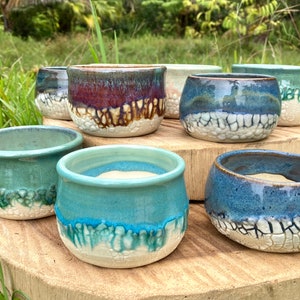 May include: A collection of seven small ceramic bowls with rounded shapes, featuring a two-tone design with smooth, glossy colored glazes on the upper sections and textured, white, crackled surfaces on the lower portions in various blue and green hues.