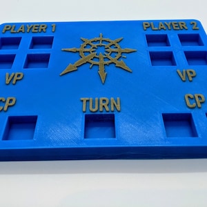May include: A blue game board with slots for Player 1 and Player 2. The board has text that reads "VP", "CP", and "TURN". A gold-colored emblem is in the center. This board is designed for tabletop gaming.