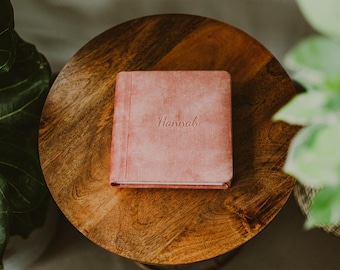 May include: A square, dusty rose-colored album with a textured cover. The name Hannah is embossed in a cursive script in the center of the front cover. The album features a vertical stitched detail along the left side of the front cover.