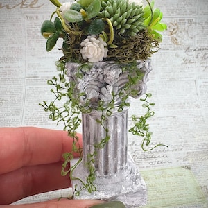 May include: Miniature gray and white column with a floral arrangement on top. The arrangement includes green succulents, white flowers, and moss. Green vines cascade down the column. The text "TYPEWRITER" and "YOUR" are visible in the background.