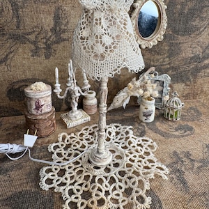 Puede incluir: Una lámpara de mesa de estilo vintage con una pantalla y base de encaje color crema. La lámpara está rodeada de otros objetos decorativos antiguos, como un pequeño espejo, un candelabro y una caja decorativa. La lámpara está enchufada a una toma de corriente.