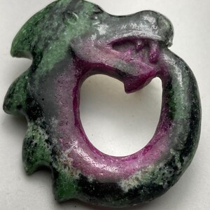 May include: A carved stone pendant in the shape of a dragon, featuring a loop design. The dragon's head is detailed with visible eyes and mouth. The stone has a mix of green, black, and deep pink hues, creating a unique and natural appearance.