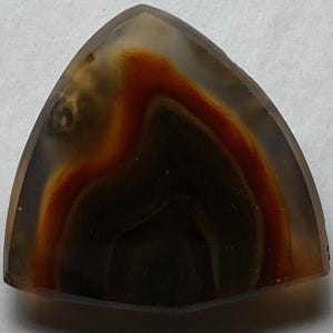 May include: A triangular-shaped gemstone with a polished surface. The stone features a dark brown base with swirling patterns of orange and tan, creating a layered effect. The edges are smooth and rounded.