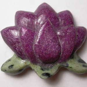 Puede incluir: Un colgante de flor de loto rosa tallada en piedra preciosa de zoisita rubí. La flor tiene una base verde con motas negras.