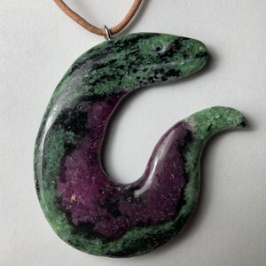 May include: A pendant necklace with a polished stone in green, black, and purple. The pendant is shaped like a stylized snake, suspended from a brown leather cord. The stone has a marbled appearance. The necklace is suitable for everyday wear.