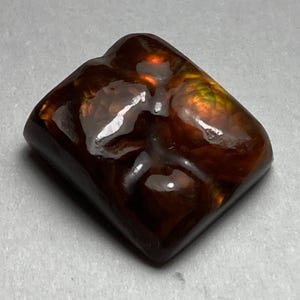 May include: A square-shaped, brown agate gemstone with red and green inclusions. The gemstone has a smooth, polished surface.