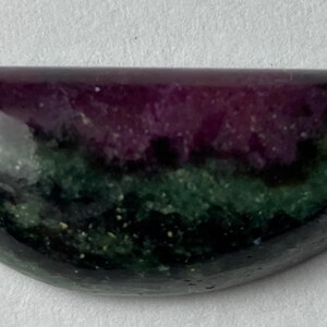 May include: A polished, semi-circular gemstone displaying a colour gradient. The top section is a deep purple, fading into green and black at the base. The stone has a smooth, glossy surface.