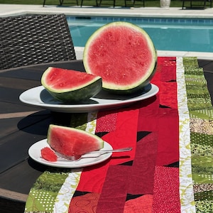 Może przedstawiać: Połówka arbuza i dwa plastry leżą na białych talerzach na czerwono-zielonym, pikowanym bieżniku. Bieżnik ma różne czerwone wzory i zielone kwiatowe wzory. Widelec leży na jednym talerzu. Scena rozgrywa się na zewnątrz w pobliżu basenu.