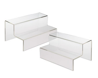 May include: Two clear acrylic display risers, each featuring a two-tier stepped design. These transparent stands are constructed with smooth, rigid panels arranged in a staircase configuration, providing elevated surfaces for organizing items.