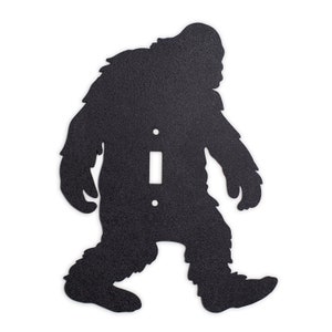 May include: Black metal light switch cover featuring a silhouette of Bigfoot walking. The switch cover is designed to fit a standard light switch.