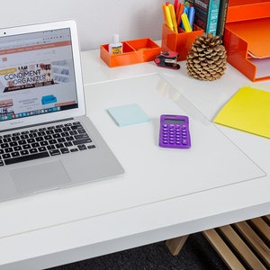 May include: A clear rectangular desk mat made of smooth plastic, positioned on a white desk surface. The mat sits beneath a silver laptop, a purple calculator, a light blue sticky note, an orange organizer with pens, and a pinecone.