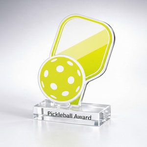 May include: A clear acrylic Pickleball Award trophy. The trophy features a pickleball paddle shape with a yellow and white design, and a yellow ball with white dots. The base of the trophy is rectangular and has the words "Pickleball Award" printed on it.