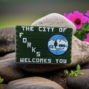 May include: A green embroidered patch with the text "THE CITY OF FORKS WELCOMES YOU." The patch features a circular emblem with a mountain, trees, and water scene in blue and white. The patch is set against a natural background of rocks and flowers.