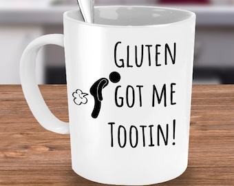 May include: White ceramic coffee mug with a black silhouette of a person with a speech bubble that says "Gluten got me tootin!"