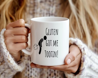 May include: White ceramic mug with the text "Gluten Got Me Tootin!" in black. The mug features a stick figure with a cloud graphic. The mug is held by a person wearing a white sweater.