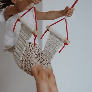 Op de afbeelding: Een jong meisje zwaait op een witte touwschommel met rode touwen. De schommel is gemaakt van een geweven materiaal en heeft houten latten aan de bovenkant.