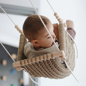 Peut inclure: Une balançoire en crochet beige avec des chevilles en bois et des cordes blanches, un jeune enfant est assis dans la balançoire.