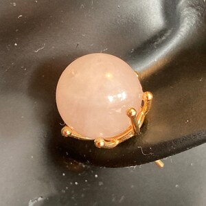 Op de afbeelding: Een close-up van een rozenkwarts oorbel. De lichtroze, bolvormige steen wordt op zijn plaats gehouden door een goudkleurige zetting. De oorbelpen is zichtbaar.