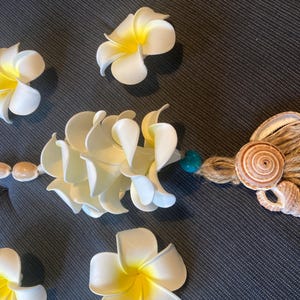 Puede incluir: Arreglo decorativo con flores de plumeria blancas y amarillas, conchas marinas y una guirnalda de conchas. La composición incluye una guirnalda central y flores sueltas, sobre una superficie texturizada gris oscuro.