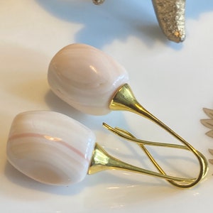 May include: A pair of earrings with light pink and white oval stones and gold-coloured metal details. The earrings have a distinctive pin-like design. The background is white, with a gold-coloured starfish and other decorative elements.