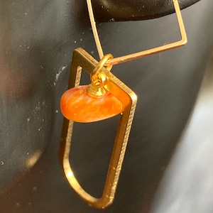 Puede incluir: Pendientes geométricos dorados. Cada pendiente presenta un cuadrado y un triángulo conectados por una barra. Una pequeña piedra naranja en forma de lágrima cuelga del triángulo. Los pendientes se muestran sobre un fondo oscuro.