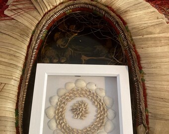 Può includere: Un'opera d'arte incorniciata con una disposizione circolare di conchiglie in una cornice bianca. Le conchiglie sono beige e bianche, creando un motivo a contrasto. L'opera d'arte è ambientata sullo sfondo di un copricapo tradizionale.
