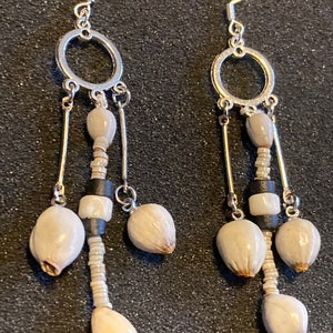 Puede incluir: Un par de pendientes colgantes con ganchos y detalles circulares plateados. Los pendientes presentan una combinación de cuentas blanquecinas, negras y beige, con cuentas más grandes en forma de lágrima en la parte inferior. Estética natural y terrosa.