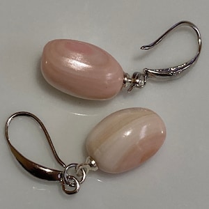May include: A pair of earrings with oval, pale pink beads and a pearlescent finish. The beads are attached to silver-coloured leverback earring findings. The earrings are displayed on a white surface.