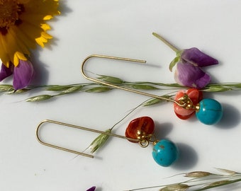 May include: A pair of gold-toned earrings with a hook design. Each earring features a turquoise bead, a cluster of coral-colored beads, and a reddish-orange stone. The earrings are displayed with flowers and greenery.