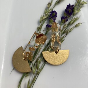 May include: A pair of gold-colored earrings with a half-circle design. The earrings have a clear, rectangular top section filled with gold flakes. The earrings are displayed on a white surface with sprigs of lavender and green grass.