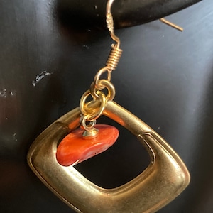 Puede incluir: Pendiente dorado con un diseño geométrico. El pendiente presenta un marco en forma de cuadrado con una piedra rojo-naranja suspendida en el centro. El pendiente tiene un cierre de gancho.