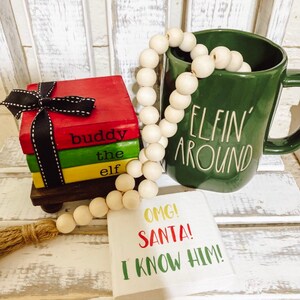 May include: A green ceramic mug with the text "Elfin' Around" on it. A wooden bead garland is wrapped around the mug. A stack of three small wooden books with the text "Buddy the Elf" on them. A white sign with the text "OMG! Santa! I know him!"